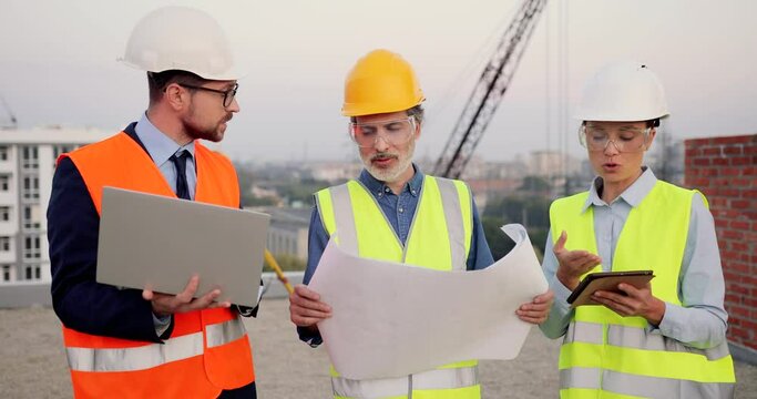 Caucasian males and female builders, architector and engineers with draft plan of building and tablet device talking on constructing site. Architector and investor, men and woman constructors working.