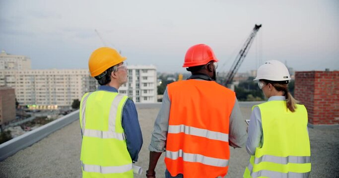 Rear. Mixed-races male and female builders in casques walking at roof top of building construction and talking. Back view on multi ethnic foreman and workers working and discussing constructing.