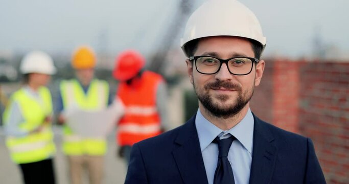 Portrait of handsome Caucasian young man architector in casque, suit, tie and glasses at construction site looking straight. Male investor at building. Mixed-races constructors talking on background.