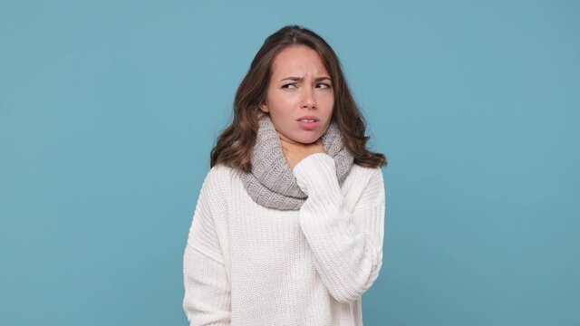 Tired displeased young brunette woman in sweater gray scarf isolated on pastel blue background studio. Healthy lifestyle ill sick treatment cold season concept. Coughing put hand on throat freezing