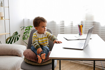 Two year old boy staring at the laptop screen with fascinated facial expression. Toddler boy sitting by the table at home with notebook computer. Close up, copy space, background.