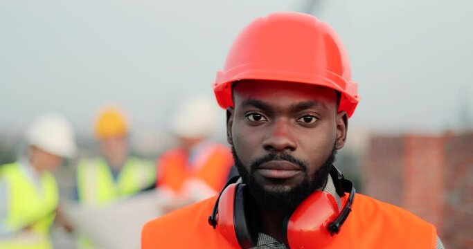 Portrait of handsome African American young man in casque and headphones turning to camera at construction site and looking at straight. Good looking male builder at building outdoor Construct concept