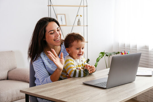 Young beautiful woman & her two year old son sitting by the desk at home using laptop for a video call. Mother & toddler child waving at the screen during video chat. Close up, copy space, background,