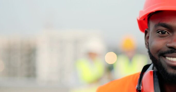 Portrait of half face of handsome African American young man in casque and headphones looking at camera at construction site and smiling. Close up of happy male builder at building. Constructing.