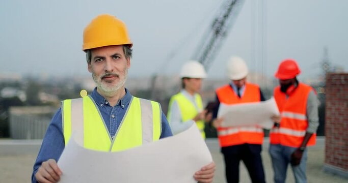 Portrait of handsome senior Caucasian man in casque hoding construction draft plan and looking a camera. Good looking male builder with beard at building outdoors. Construct concept.