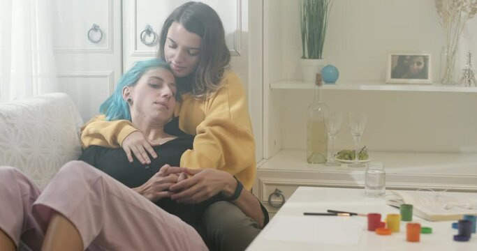 Young girlfriends resting on sofa at home. Young woman hugging and touching hand of girlfriend with dyed hair while relaxing on couch in cozy living room at home