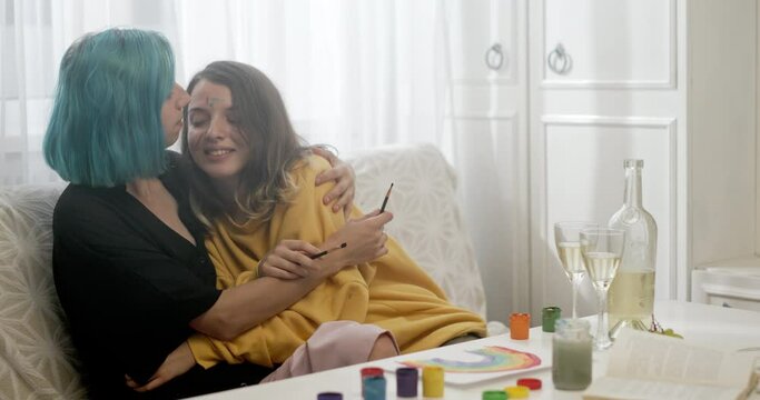Happy girlfriends hugging and kissing while painting. Cheerful young women embracing each other and kissing with smile while sitting on sofa and painting rainbow at home