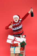 Smiling senior man in santa hat holding glass and bottle of champagne near presents in shopping cart isolated on red