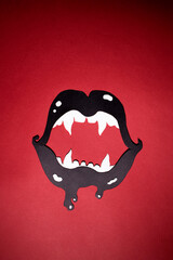 Black lips with white fangs cut out of paper on a red background. The vertical photo of paper art...
