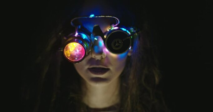 Woman in grungy VR glasses in dark room. Mad female scientist in handmade invented VR goggles glowing with light in dark experiencing virtual reality