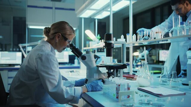 Medical Research Scientist Looks at Biological Samples Under Digital Microscope in Applied Science Laboratory. Beautiful Caucasian Lab Engineer in White Coat Working on Vaccine and Medicine.