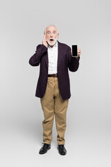 Shocked elderly man with hand near head holding smartphone with blank screen on grey background