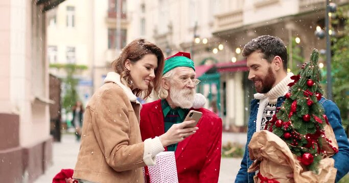 Portrait of cheerful Caucasian female and male couple standing outdoor with Santa Claus and taking selfie photo on street. Joyful family with Santa take pictures on cellphone. Christmas concept