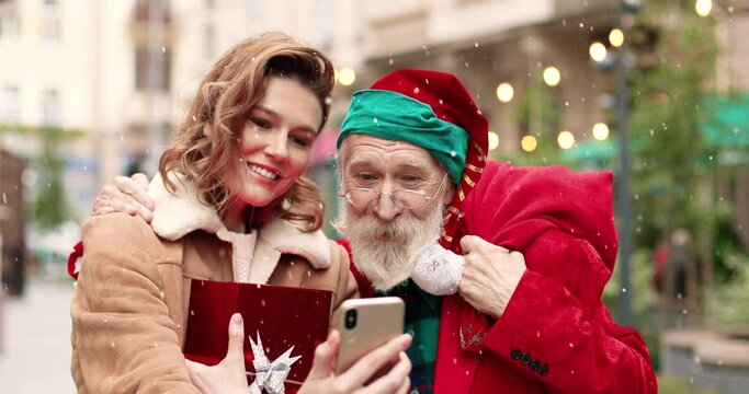 Close up portrait of happy beautiful woman taking selfie photos on smartphone with senior Caucasian man dressed like Santa Clause while standing in decorated city on festive day. Xmas concept