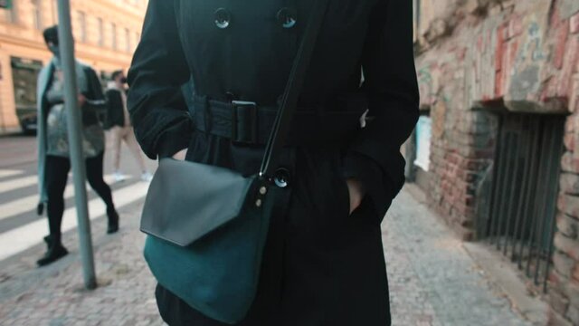 Urban autumn fashion is shown on a hurrying moving young business woman. Women's double-breasted coat, handbag. A black color fabric mask on face prevents droplets from spreading. Hands in pockets.