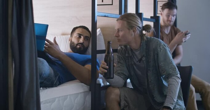 Young Caucasian man drinking beer in inn or hostel and talking with Middle Eastern friend lying on bed and surfing Internet on laptop. Relaxed fellows chatting at bedtime. Cinema 4k ProRes HQ.