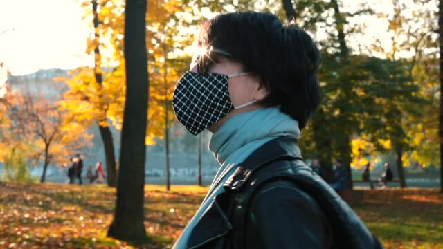 A woman wearing a mask puts glasses on her eyes. Walk in the city park in autumn on a sunny day. Eye protection from rays of bright sun. On background are yellow trees.