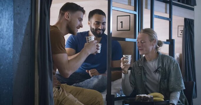 Three positive multiethnic friends celebrating holiday in hostel or inn. Portrait of happy relaxed Caucasian and Middle Eastern men clinking paper cups and talking. Cinema 4k ProRes HQ.