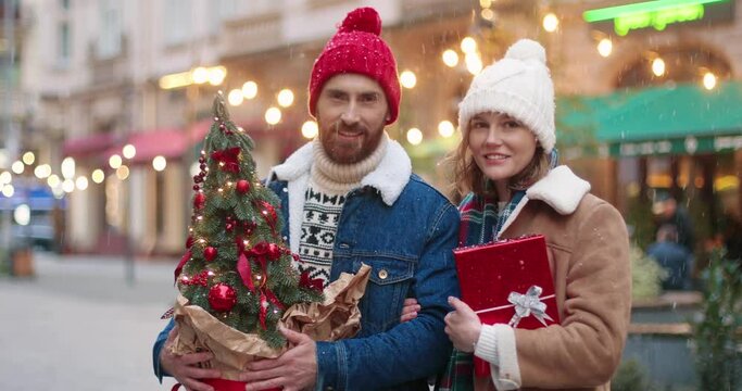 Joyful couple standing in snowy town and smiling to camera on christmas holiday. Lovely cute Caucasian family with xmas presents outdoor. Romantic relations concept. Close up portrait