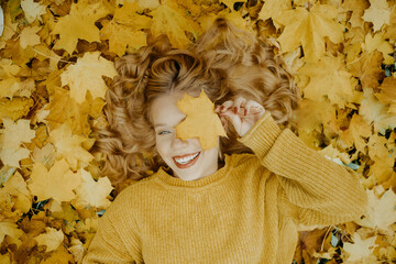 Beautiful girl lies in the park in autumn. A teenager in a yellow sweater on a background of orange leaves. The blonde smiles and laughs. Fashion and Style. Woman covering eyes with leaf and smiling