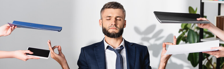 Businessman meditating near colleagues with smartphone and paper folders in office, banner