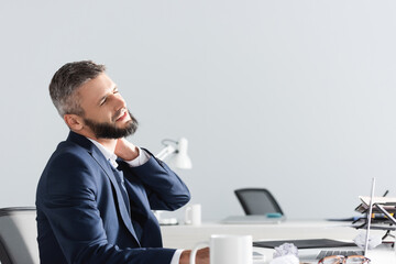 Businessman feeling neck pain near laptop and clumped paper on blurred foreground in office
