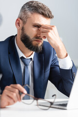 Exhausted businessman in suit holding eyeglasses while touching eyes near laptop on blurred foreground