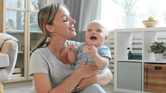 Medium shot of happy young Caucasian woman wearing casual clothes sitting on floor in living room with laughing blond-haired little son in her hands