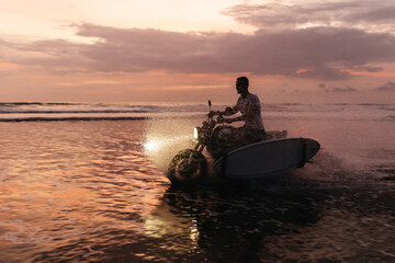 Surfer rides on motorbike with surfboard at sunset ocean beach.