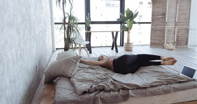 Young woman lies in bed doing exercises in morning lazy sports at home in horizontal position swinging legs from side to side on sheet stretching muscles of lower back practices yoga asanas for spine