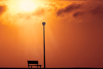 A Wooden bench and street light with a background view of a red sunset on sunset and a bright sun