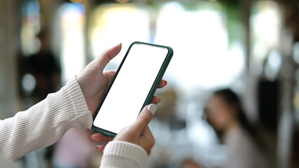 Cropped shot woman in white sweater hands holding blank screen smartphone over blurred interior of meeting room background.
