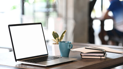 Mockup blank screen laptop, coffee cup, plant and notebook on desk in office room background.