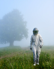 Cosmonaut in space suit strolling down the foggy meadow. Space traveler in helmet walking on green grass near the road with misty tree on background. Concept of astronautics, exploration and nature.