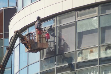 Photo of city works. A worker washes the facade of a building using a car platform, a lifting mechanism. Iron, glass, and concrete, nothing extra.