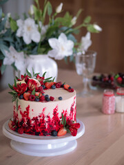 Big beautiful red velvet cake, with flowers and berries on top. Dessert on the kitchen table.