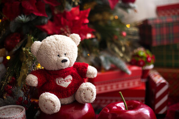 White Teddy bear in red knitted sweater with heart on the chest and the words Love near Christmas tree among the gift boxes. Gift to your beloved, Declaration of love, poinsettia decor. Copy space