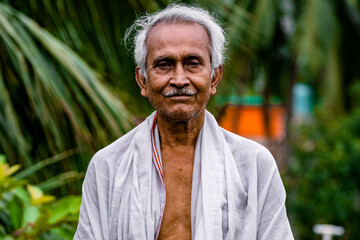 Close-up Portrait of Indian Senior Man Standing Alone. Happy old man, wearing white dress, giving pose with style and smiling at the camera. Elderly Man Is Enjoying Retirement In Nature In Afternoon.