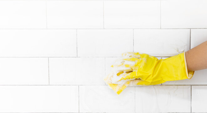 A female hand in a yellow latex glove sponges a tile. There is a lot of foam in the sink. The concept of cleaning the bathroom or kitchen, cleanliness. Copyspace.