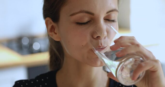 Close up head shot healthy happy positive young attractive woman making sips, drinking stilled pure mineral water from glass, normalizing bowel function, feeling good at home, daily healthcare habit.