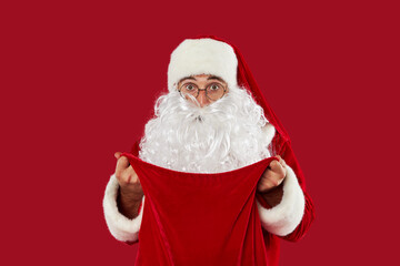 Surprised Santa Claus in round glasses peeks into a bag full of gifts on a red studio background