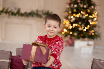 Obraz na płótnie Canvas Portrait of happy smiling little boy in red knitted sweater with Christmas present box near Christmas tree. Happy childhood. Waiting for a Christmas tale