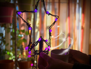 Naklejka premium Hands Of A Woman Drinking A Cup Of Coffee At Christmas. Hands Holding Christmas Lights.