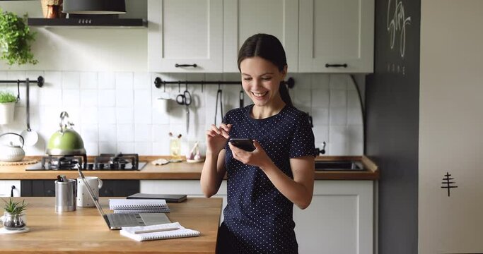 Distracted from remote job or study young pleasant happy woman using smartphone, receiving message with win notification, celebrating personal success achievement, getting paid on banking balance.