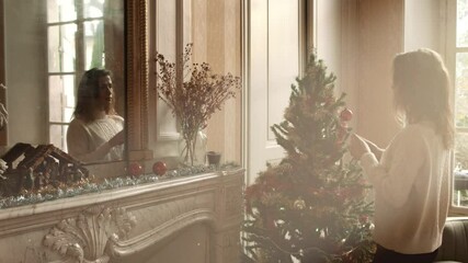 A woman decorates a Christmas tree with decorations next to a fireplace, under the sunlight from the window. Beautiful christmas scene