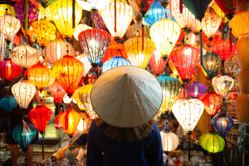 Tourist woman is wearing Non La (Vietnamese tradition hat) and looking colorful lanterns spread light on the old street of Hoi An Ancient Town - UNESCO World Heritage village.