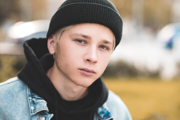 Stylish teen boy 15-16 year old wearing black knitted hat and hoodie outdoors close up. Looking at camera. Autumn season. Teenagerhood.