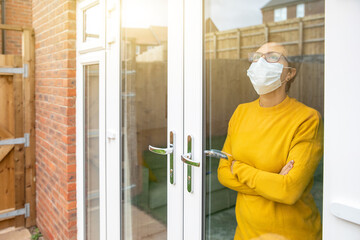 Woman wearing a face mask isolating at home and looking out of the window - Young woman feeling sad alone at home due to coronavirus quarantine and lockdown