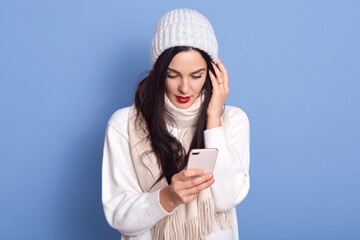 Photo of stylish pretty lady looks at telephone screen, chatting messages, wear warm casual hat, scarf, pullover, posing isolated over blue background, sees something astonishing at mobile screen.