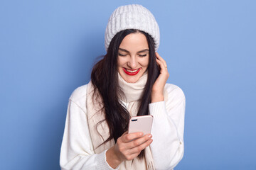 Portrait of nice attractive satisfied cheerful girl holding in hands and using cell phone, lady stands isolated over bright blue background, female wearing white pullover, scarf and cap.